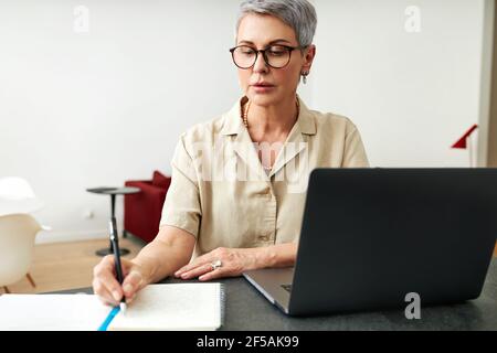 Mature woman writing on a note pad while using laptop computer at home Stock Photo
