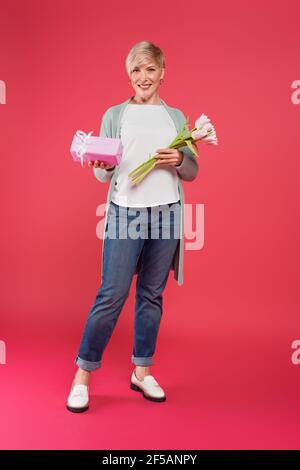 full length view of trendy middle aged woman posing with tulips and gift box on pink Stock Photo