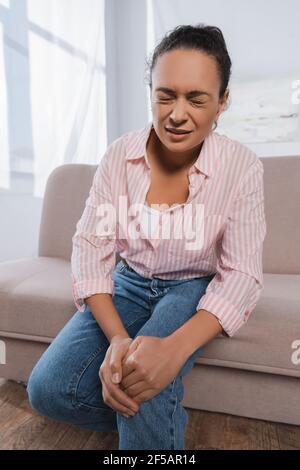 african american woman suffering from pain in knee while sitting on couch Stock Photo