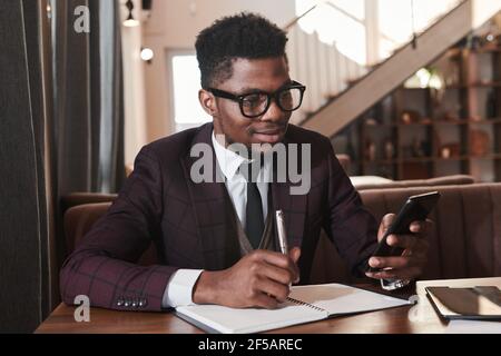 African young man in eyeglasses working online on his mobile phone and making notes in notebook sitting in the restaurant Stock Photo
