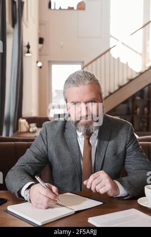 Portrait of mature bearded businessman looking at camera while writing plans in notebook in the restaurant Stock Photo