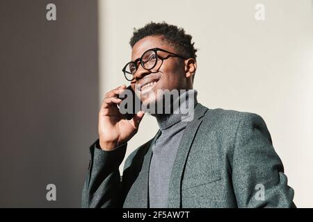 African young businessman in eyeglasses smiling while talking on mobile phone outdoors Stock Photo