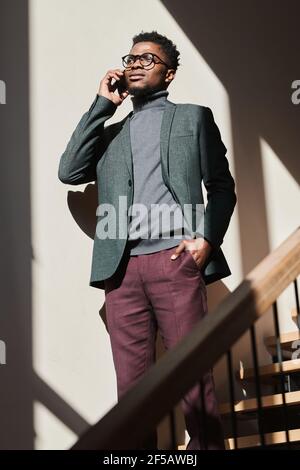 African young businessman in casual clothing standing on stairs and talking on mobile phone Stock Photo