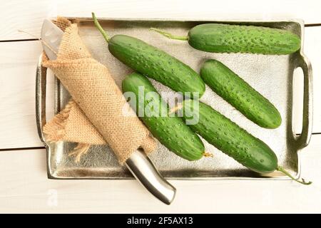 Several ripe dark green organic delicious cucumbers on a metal tray with a metal knife wrapped in a jute napkin, on a white wood counter top. Stock Photo