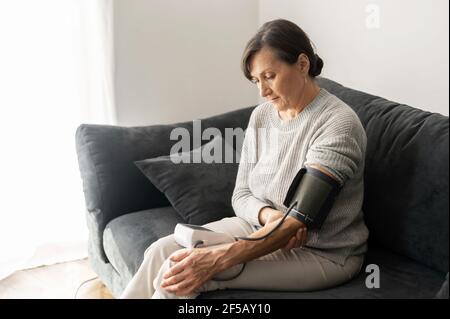 Senior woman checking blood pressure level at home, older female suffering from high blood pressure sitting at a couch and using a pulsometer, tonometer Stock Photo