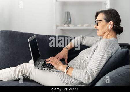 Contemporary senior woman enjoys chatting in social networks, lying on the comfortable sofa with a laptop, spends leisure time in network, texting email, connect with friends on the distance Stock Photo