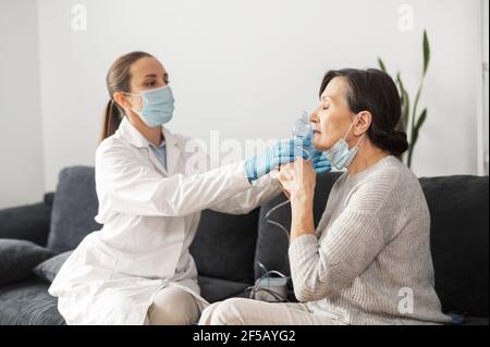 Female doctor wearing a white gown and a face mask, putting oxygen mask on the face of a senior woman, nurse cares female patient at home during pandemic Stock Photo