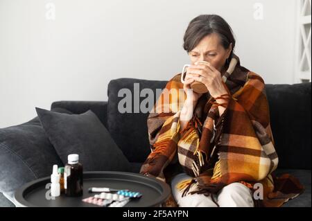 A senior woman feels unwell, sits down on the couch alone at home, covered with blanket feels chill, an elderly woman suffers from viral disease, has fever, treats herself with hot remedy drinks Stock Photo