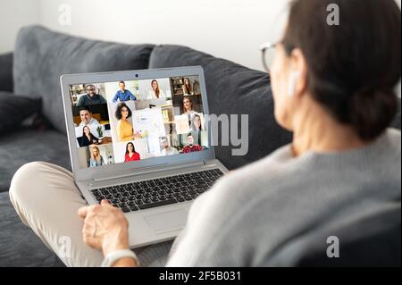 Contemporary senior woman lying on the comfortable sofa and using laptop for video connection with group of diverse people, back view an older female involved webinar, video conference Stock Photo