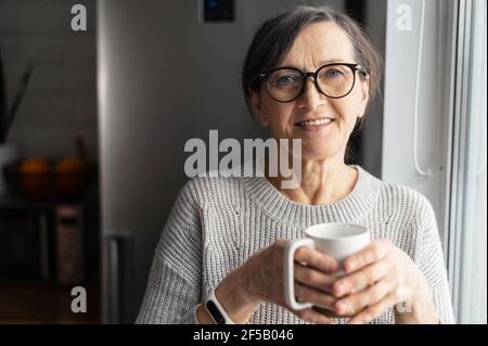 Charming elderly woman wearing stylish eyeglasses stands with cup of morning coffee looks at the camera and smiles, friendly female with mug of hot drink spends time at home Stock Photo
