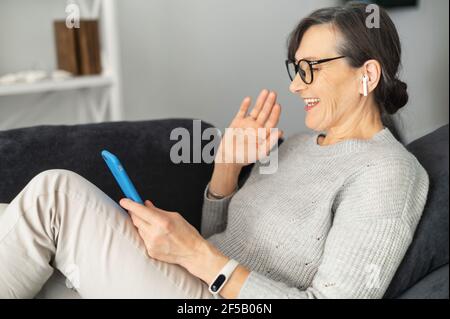 Happy senior woman making video call on the smartphone, a mature female lying down on the couch, holds mobile phone and waving hello into webcam, greeting family or friends on the distance Stock Photo