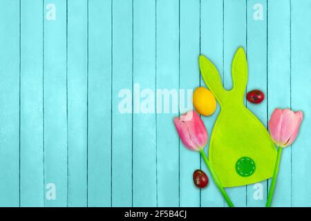Green Easter bunny with painted spotted and yellow eggs and pink tulips on turquoise planks. Easter background. Copy space Stock Photo