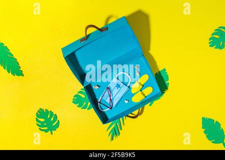 Travelling during corona virus epidemic concept. Protective mask, sun glasses and flip flops in blue suitcase on yellow Stock Photo