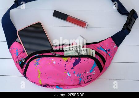 Banana bag with phone, lipstick, cream and dollar bills. Waist bag pink color on a white wood background. A small fabric bag with two compartments for Stock Photo