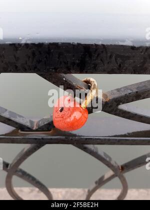 Red lock in the shape of a heart on the metal fence of the city embankment. The lock is metal hinged. Stock Photo