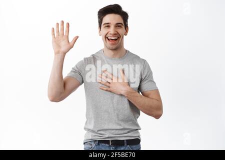 Happy smiling adult man introduce himself, raising hand and hold palm on chest, swearing, making an oath or promise, standing cheerful against white Stock Photo