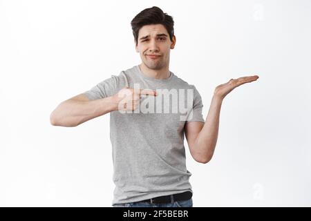 Skeptical guy pointing at open hand and look disappointed, holding copy space in hand, complaining, standing over white background Stock Photo