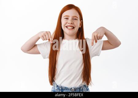 Cute redhead little girl with freckles showing promotional text, pointing down at banner and smiling toothy. Ginger child making announcement Stock Photo