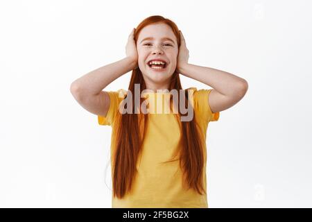 Happy little redhead girl with freckles laughing, smiling and covering ears with hands, unwilling to listen, playing around, standing over white Stock Photo