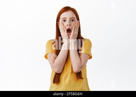 Surprised redhead girl with freckles gasping, drop jaw and stare impressed at camera, holding hands on face startled and amazed, white background Stock Photo
