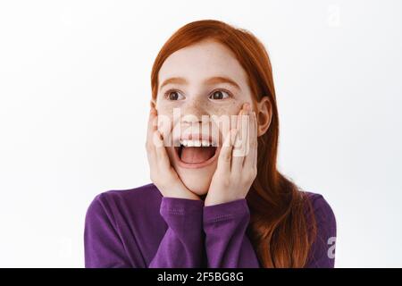 Little redhead girl look surprised and happy aside, amazed with super cool thing, smiling impressed or fascinated, stare with admiration at banner Stock Photo