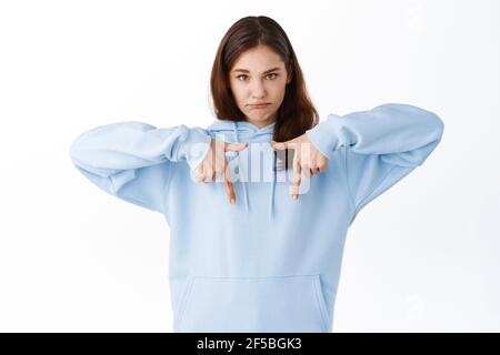 Young upset girl with gloomy sad face, pointing fingers down at bottom copy space, complaining about unfair situation, standing displeased against Stock Photo