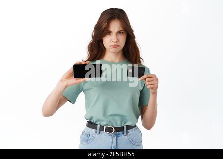 Sad and gloomy girl frowning upset, showing horizontal empty mobile screen and plastic credit card, complaining, standing against white background Stock Photo