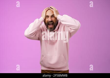 Outraged furious mad adult man bearded yelling shocked despair hatefully looking camera insane holding hands head frowning shouting losing bet Stock Photo