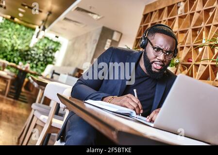 Serious businessman with headphones sitting in cafe in front of laptop and networking Stock Photo