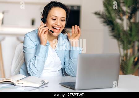 Confident attractive mixed race woman wearing headphones, freelancer, manager or real estate agent working remotely, talks by conference call with colleague or client, gesturing hand, smiling friendly Stock Photo