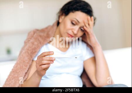 Sick tired upset adult pregnant mixed race woman sitting on sofa in living room under blanket supporting her head with hand while taking temperature with thermometer, she has cold, flu Stock Photo