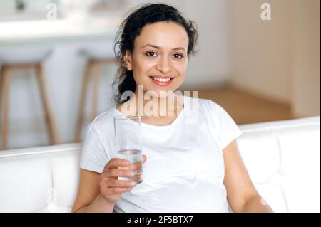 Beautiful happy mixed race woman sitting on sofa in living room, holding glass of clean water, drinking daily water intake, living a healthy lifestyle, looking at camera, smiling friendly Stock Photo