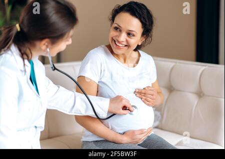 Happy attractive adult mixed race woman, sitting on sofa hugging belly, at home or hospital at doctor's appointment, friendly doctor is listening to baby with stethoscope, checking child health, smile Stock Photo