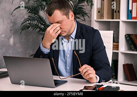 Eye fatigue, poor eyesight, office work. The young man takes off his glasses and rubs his eyes with his hand sitting at a table with a laptop in a sui Stock Photo