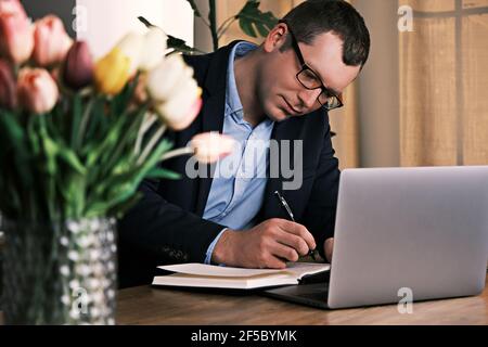 Businessman wearing suit and glasses is writing something in a notebook sitting behind a laptop. Selective focus. Bouquet of tulips in the foreground Stock Photo