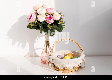 Easter colorful eggs in busket on white wooden table. Bouquet of rose flowers on background. Happy Easter. Springtime mood. Stock Photo