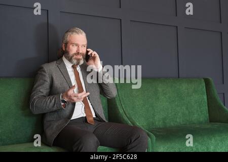 Senior bearded businessman in elegant suit talking on mobile phone with colleague sitting on comfortable sofa at office Stock Photo