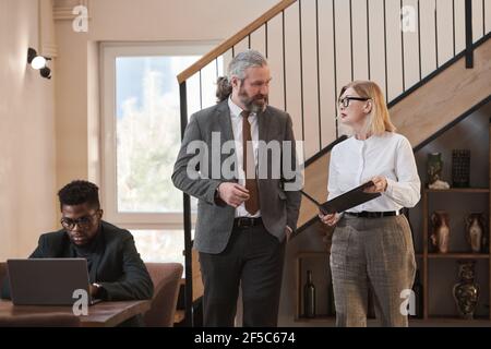 Mature business couple talking to each other and discussing business documents while walking along the office Stock Photo