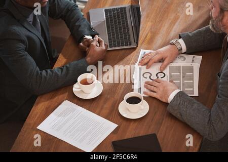 Close-up of business partners sitting at the table with cups of coffee and working with documents using laptop Stock Photo