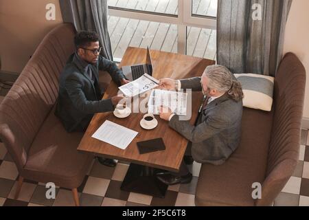 Two businessmen working at the table with laptop and documents and discussing teamwork during a meeting in restaurant Stock Photo