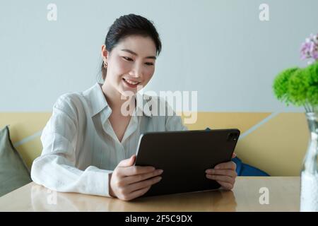Smiling young asian woman entrepreneur checking order by digital tablet while sitting at her home office. Work From Home concept. Stock Photo