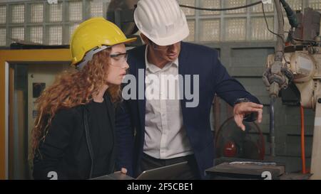 Caucasian maintenance engineer man and diverse female factory workers in hardhats talking and present or explain robot welding machine on plant, Manag Stock Photo