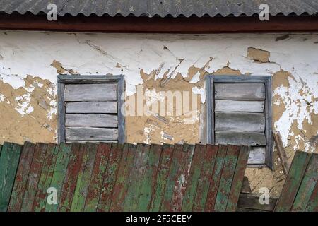 Facade of abandoned crumbling mud house in a village with boarded up windows behind a green wooden fence. Close-up. Outdoors. Stock Photo