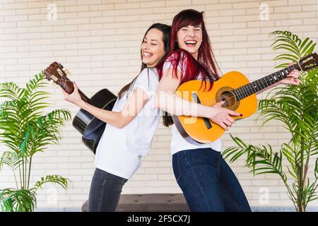 Side view of young women smiling and playing Spanish guitars while standing back to back against brick wall Stock Photo