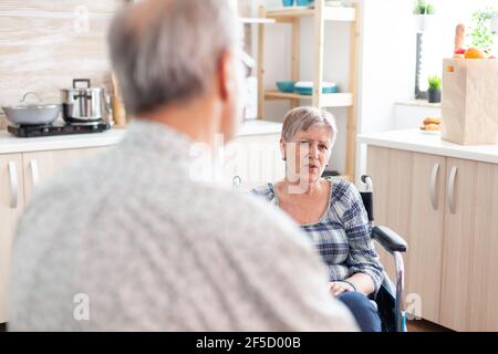 Worried disabled senior woman in wheelchair talking with husband sitting in kitchen. Old couple having a conversation about treatment, man living with disabled person with walking disabilities Stock Photo