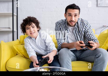 KYIV, UKRAINE - JANUARY 15, 2021: concentrated muslim dad and son gaming with joysticks at home Stock Photo