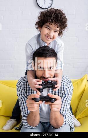 KYIV, UKRAINE - JANUARY 15, 2021: cheerful arabian boy playing video game with father at home Stock Photo