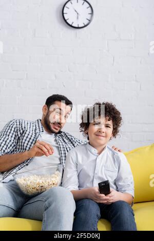 frightened arabian man eating popcorn while watching tv with son at home Stock Photo