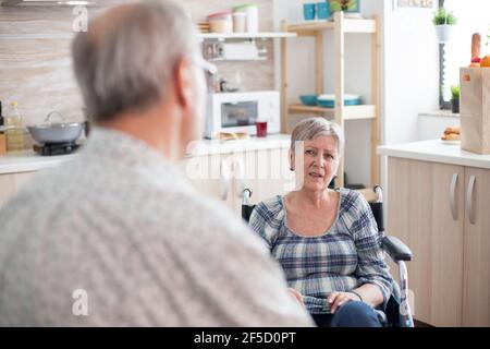 Gray Haired man chatting with paralysed wife. Retired invalid woman in wheelchair having a conversation with old elderly husband in kitchen. Old man talking with wife. Living with disabled person with walking disabilities Stock Photo