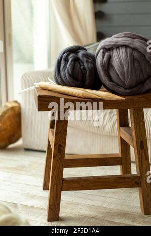 Two large balls of merino wool lying on a wooden stool with wooden knitting needles against the background of a window and a white sofa Stock Photo